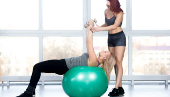 Fitness practice, two fit beautiful females working out in sports club, instructor assists blond young woman in doing exercise with dumbbells on green Swiss ball in class, full length